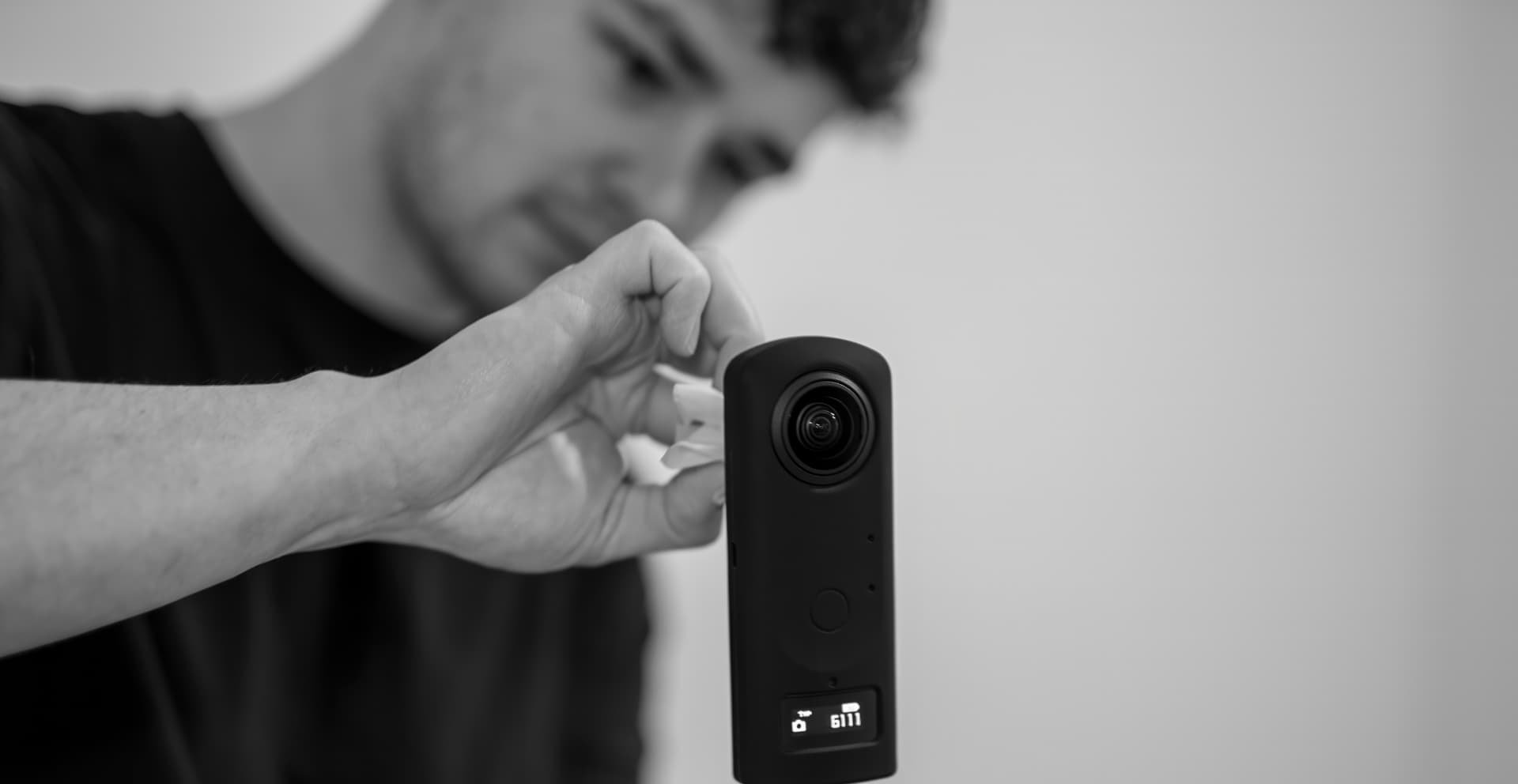 Schwarzweißaufnahme: Julian Becker reinigt das Objektiv seiner 360°-Kamera, die im Vordergrund auf einem Stativ steht